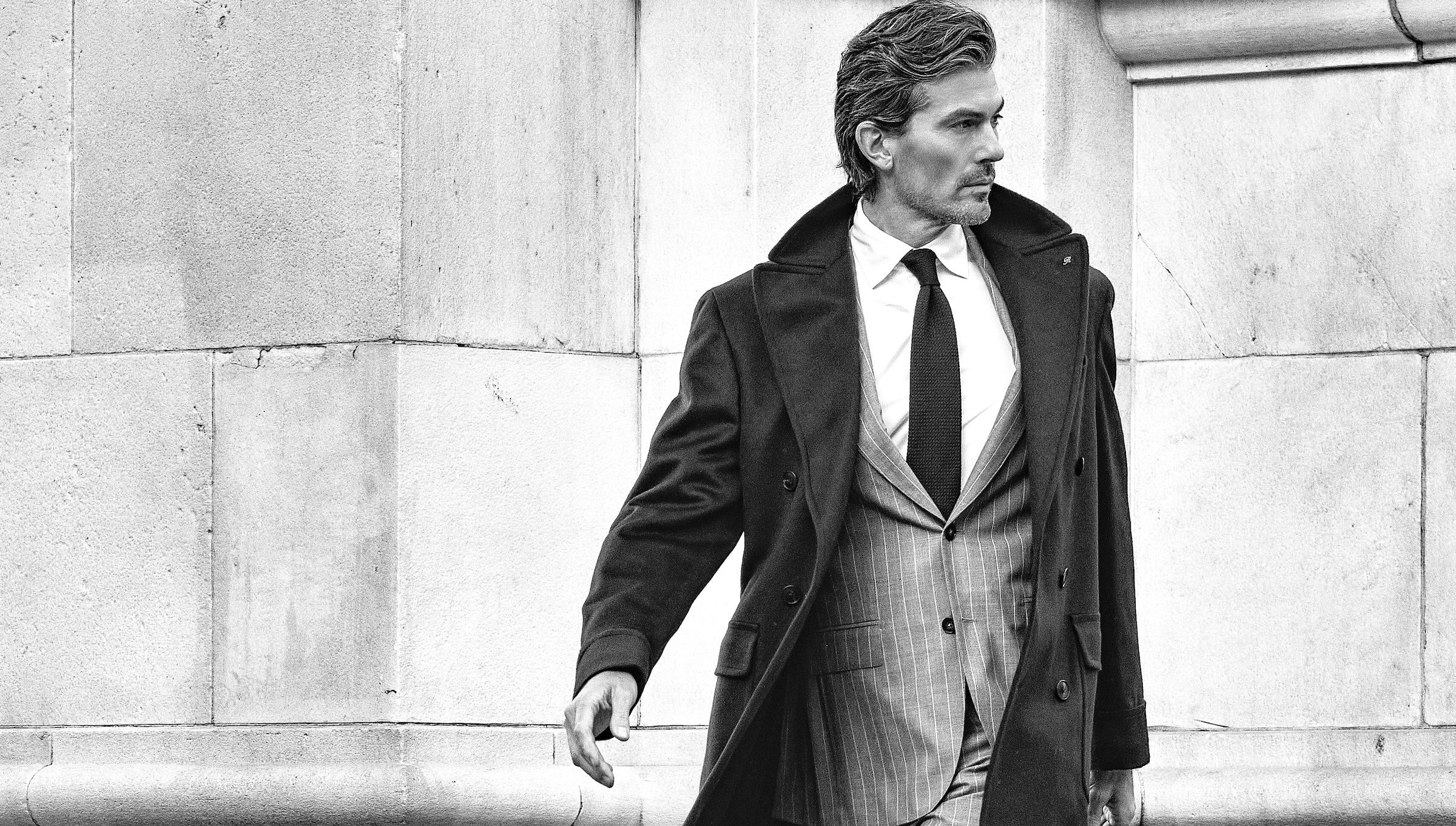 Man in a suit and overcoat standing against a stone wall.