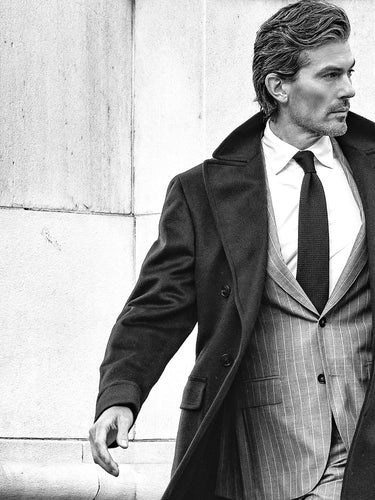 Man in a suit and overcoat standing against a stone wall.