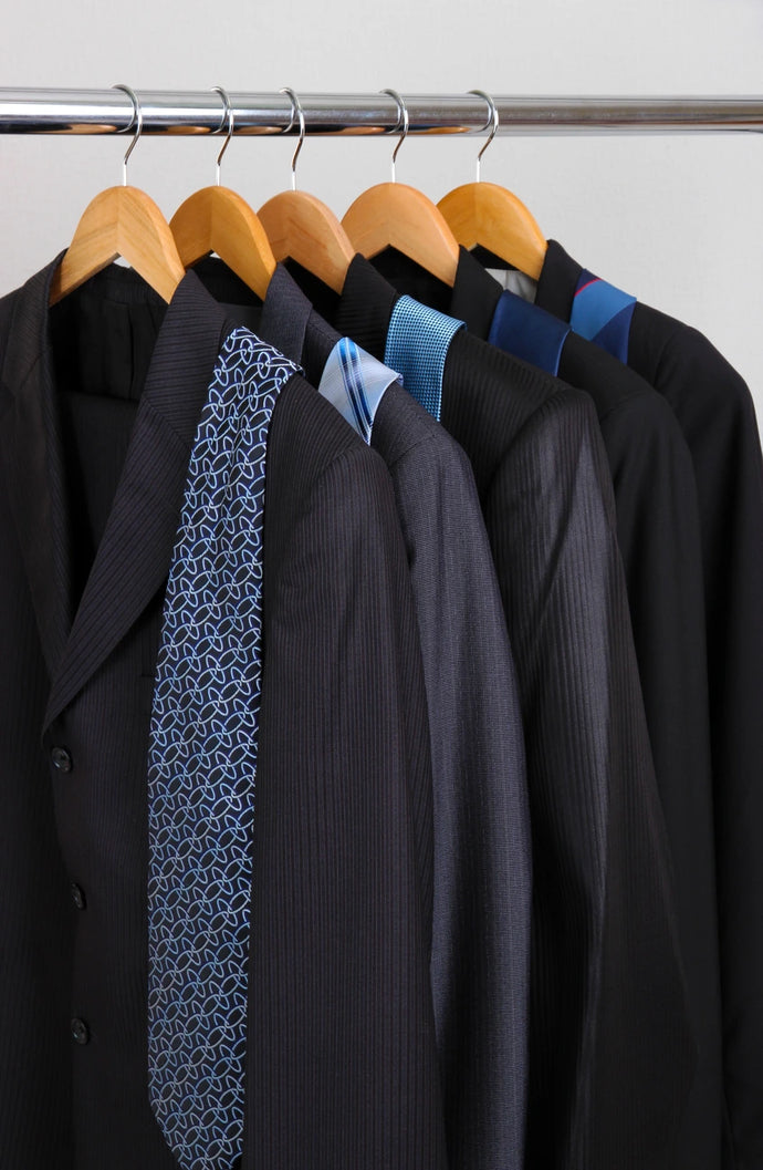 Black suits with blue patterned ties on wooden hangers against a white wall.